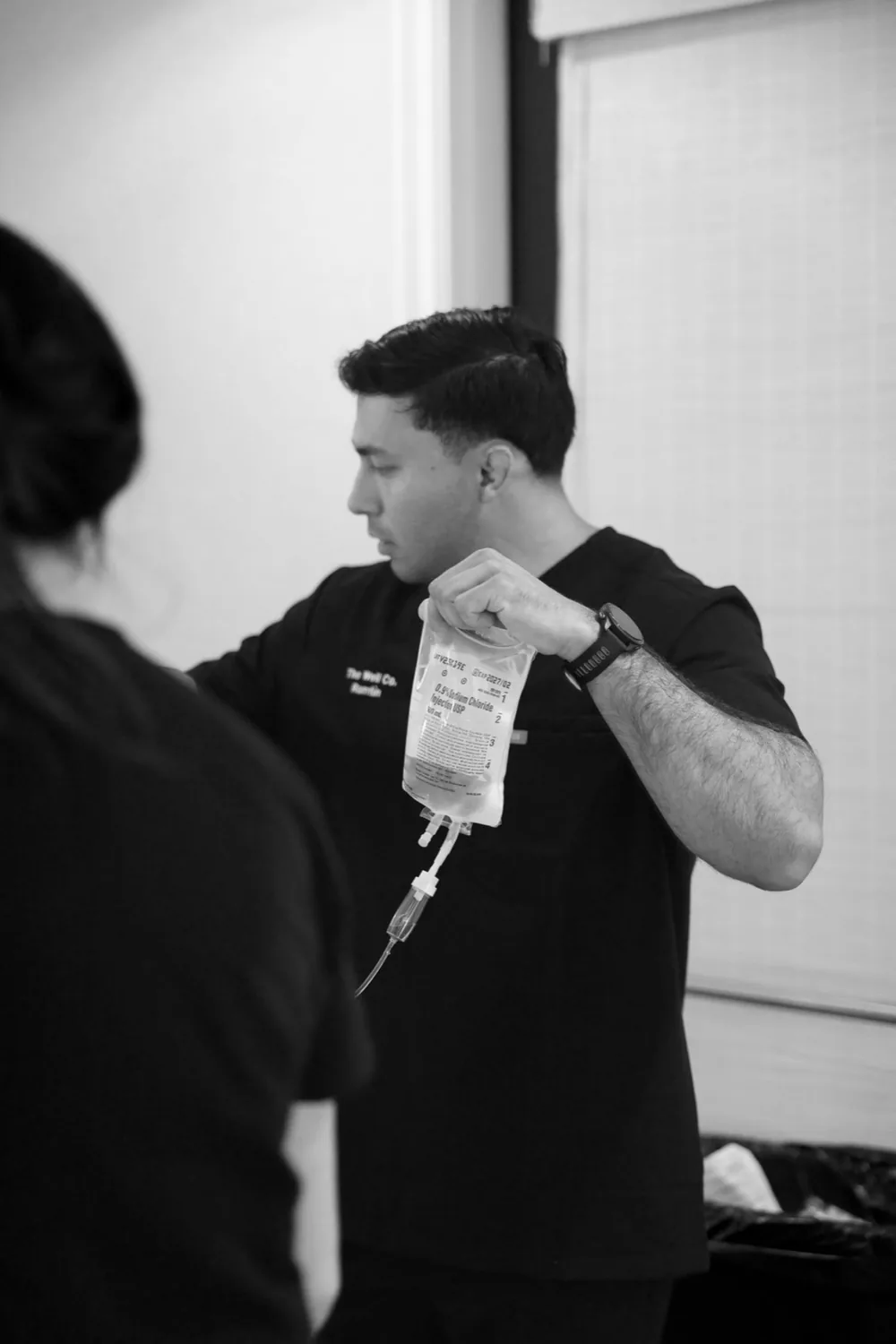 A Well Company team member in black scrubs preparing a labeled saline IV bag, in black and white